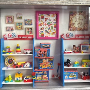 Op de afbeelding: Een ingelijste schaduwdoos met miniatuurspeelgoed en ingelijste kunstwerken. De display bevat speelgoedplanken met divers speelgoed, een roze ingelijste foto en kleine ingelijste foto's. De woorden "Classic Hits" zijn zichtbaar op de planken.