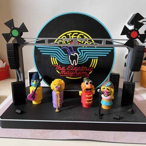 Könnte beinhalten: Eine Bühnenkulisse mit der Band The Electric Mayhem. Die Bühne hat eine schwarze Basis mit Lautsprechern, eine graue Überkopfstruktur und eine große schwarze Vinyl-Schallplatten-Kulisse mit dem Logo der Band. Die Bandmitglieder sind farbenfrohe Figuren.