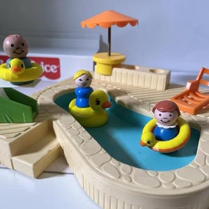 May include: A vintage Fisher-Price toy pool set with a light beige pool, blue water, and several figures. The set includes a slide, umbrella, lounge chair, and figures with duck floats. The figures are wearing blue and yellow clothing.