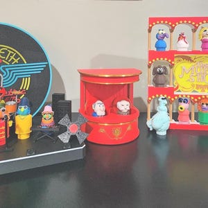 Puede incluir: Una colección de figuras de personajes de los Muppets que incluye un set de la banda Dr. Teeth and the Electric Mayhem con un logo circular, un escenario de balcón rojo con dos figuras y un estante de varios niveles con varios personajes.