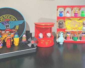 May include: A collection of Muppet character figurines including a Dr. Teeth and the Electric Mayhem band set with a circular logo backdrop, a red balcony stage with two figures, and a multi-tiered display shelf featuring various Muppet characters.