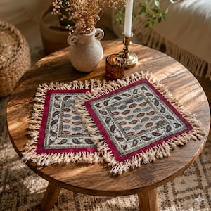 May include: Two square decorative coasters with a paisley pattern in shades of red, blue, and beige. Each coaster has a red border and is fringed with beige tassels. They are displayed on a round wooden table.