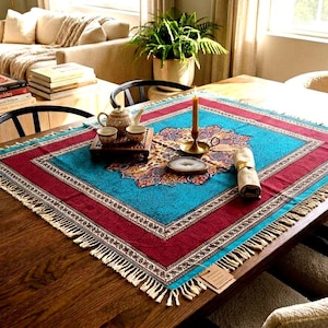 May include: A square tablecloth featuring a vibrant turquoise center with a central floral medallion, surrounded by a deep red border and intricate patterned trim. The edges are finished with a fringe, and it is displayed on a wooden table.