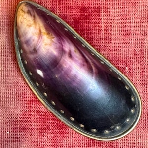 May include: An elongated, oval-shaped decorative object with a silver-colored metal trim. The interior features a swirling pattern of purple, black, and cream colors. The trim is adorned with small, evenly spaced rivets.