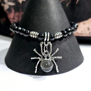 Könnte beinhalten: Ein schwarzes Perlenarmband mit einem silbernen Spinnenanhänger und einem dunkelgrauen Edelstein. Das Armband befindet sich auf einem schwarzen, kegelförmigen Ständer.