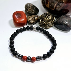 Può includere: Un braccialetto in pietra lavica nera con perline rosse e argentate. Il braccialetto è su una superficie bianca con varie pietre e un oggetto metallico decorativo.