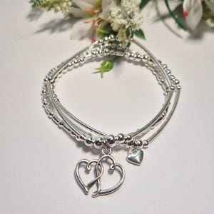 Op de afbeelding: Een meerstrengs zilverkleurige armband bestaande uit een combinatie van gladde ronde kralen en dunne metalen staafjes. De armband is versierd met een bungelende bedel bestaande uit twee in elkaar grijpende open hartjes en een klein massief hartje.