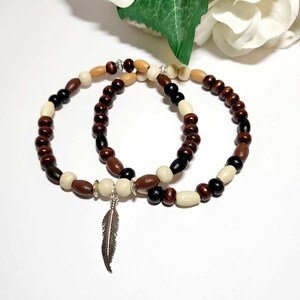Pode incluir: Duas pulseiras de contas de madeira com um fecho de pena de prata. As pulseiras são de cor castanho, preto e creme.