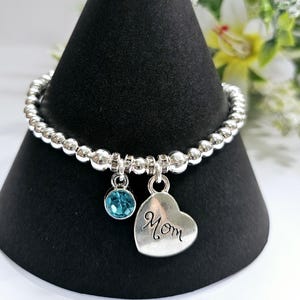 May include: A silver beaded bracelet with a turquoise crystal charm and a silver heart charm that says "Mom".