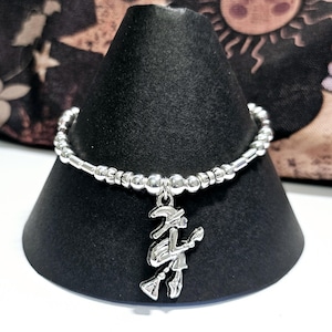 Könnte beinhalten: Ein silbernes Armband mit einem Hexen-Anhänger. Das Armband besteht aus kleinen silbernen Perlen und hat einen silbernen Anhänger einer Hexe, die auf einem Besen reitet.