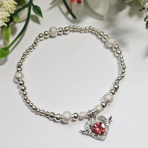 May include: Silver bracelet with a heart-shaped charm. The charm has a red gemstone centre, surrounded by clear stones and small wings. The bracelet is made of small, round silver beads and larger, textured silver beads.