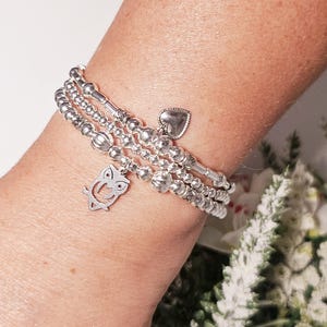Puede incluir: Un brazalete de plata con múltiples hileras de cuentas y dijes, incluyendo un búho y un corazón.