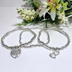 Puede incluir: Tres pulseras de cuentas plateadas con dijes. Una pulsera tiene un dije de corazón con un diseño de árbol. La otra pulsera tiene un dije de corazón doble.