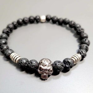 Könnte beinhalten: Schwarzes Lava-Stein-Armband mit einem silbernen Totenkopf-Anhänger.