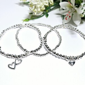 Puede incluir: Tres pulseras de cuentas plateadas con dijes de corazón. Las pulseras están apiladas una encima de la otra.