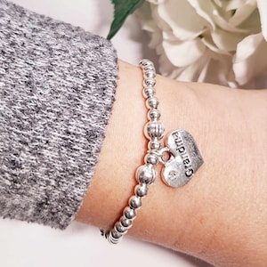 May include: Silver beaded bracelet featuring a heart-shaped charm inscribed with "Grandma". The charm includes a small crystal. The bracelet is displayed on a wrist, with a grey sleeve visible.