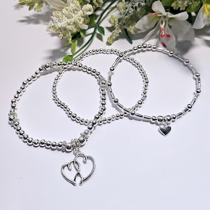 May include: Three silver beaded bracelets with heart charms. The bracelets are stacked on top of each other. The charm on the bottom bracelet is a single heart, while the charm on the top bracelet is two hearts intertwined.