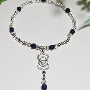 Op de afbeelding: Zilverkleurige kralenarmband met donkerblauwe kralen en een bungelende bedel. De bedel heeft een geometrisch ontwerp met een driehoek in het midden. De armband staat op een witte achtergrond.