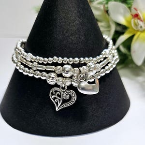 Può includere: Tre braccialetti d'argento con ciondoli a forma di cuore. Un ciondolo è un cuore pieno, l'altro è un cuore in filigrana. I braccialetti sono esposti su un supporto nero a forma di cono, con un fiore bianco e giallo sullo sfondo.