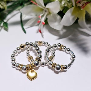 May include: Three silver and gold beaded rings with a gold heart charm. The rings are stacked on top of each other.