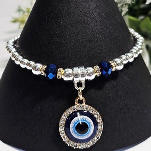 Peut inclure: Bracelet en argent avec des perles et des accents bleus et dorés. Il comporte un pendentif rond avec un motif d'œil bleu et blanc, entouré de cristaux transparents. Le bracelet est présenté sur un présentoir noir.