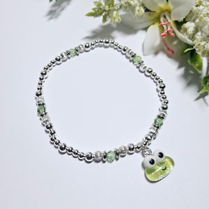 May include: A silver beaded bracelet with green crystal beads and a green frog charm. The frog has white eyes and a smiling mouth.