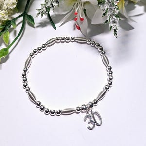 Op de afbeelding: Een zilveren armband met een bedel die "30" zegt. De armband is gemaakt van kleine zilveren kralen en heeft een delicaat ontwerp.