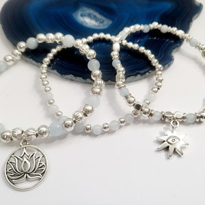 Op de afbeelding: Drie zilveren kralenarmbanden met blauwe edelsteenkralen. Een armband heeft een zilveren lotusbloembedel, en een andere heeft een zilveren sterbedel met een oog in het midden.
