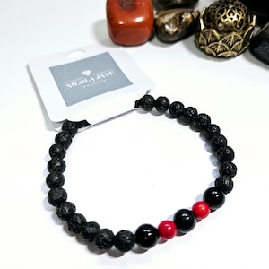 May include: Black lava rock beaded bracelet with red and black accent beads. The bracelet is on a white background with a card that says "Nicola Jane Jewellery".