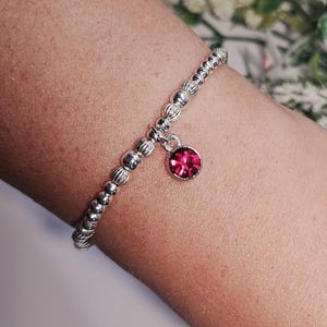 Peut inclure: Un bracelet en argent composé de perles et orné d'un petit pendentif rond en pierre rose. Le bracelet est porté sur un poignet, mettant en valeur le design délicat et la couleur vive du pendentif. Les perles du bracelet ont un aspect texturé.