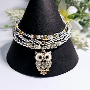 Pode incluir: Um bracelete de contas prateadas e douradas com um fecho de coruja. O fecho de coruja tem esmalte branco e um cristal transparente no centro.