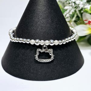 Puede incluir: Un brazalete de plata con un dije de Hello Kitty de plata. El dije está adornado con pequeños diamantes de imitación transparentes. El brazalete está hecho de pequeñas cuentas de plata.