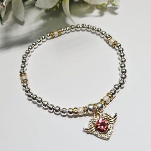 Könnte beinhalten: Silbernes Perlenarmband mit goldenen Akzenten und einem Herz-Anhänger. Der Anhänger hat einen rosa Edelstein in der Mitte, umgeben von kleinen klaren Steinen und goldenen Flügeln. Auf weißem Hintergrund.