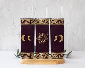 May include: Three stainless steel tumblers with a burgundy base and gold accents. The tumblers feature a sun and moon design with a decorative border. The tumblers are displayed on a wooden stand.