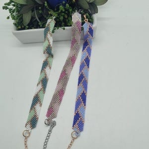 Könnte beinhalten: Drei Armbänder mit Perlen in einem Zickzackmuster in Grün-, Rosa- und Blautönen. Jedes Armband hat eine Kette und einen Verschluss. Im Hintergrund befindet sich eine weiße rechteckige Vase mit einer blauen Rose und Grünzeug.