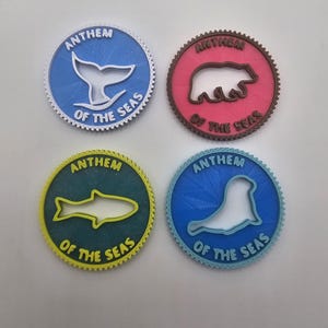 Peut inclure: Quatre dessous de verre ronds et colorés avec des motifs de la vie marine. Chaque dessous de verre présente le texte "ANTHEM OF THE SEAS" et une créature marine différente : une queue de baleine, un ours, un poisson et un phoque. Les dessous de verre sont bleus, rouges, verts et bleus.
