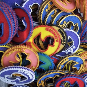 May include: A collection of colorful, 3D-printed Disney-themed medallions. The medallions feature various colors, including red, blue, yellow, and pink, with intricate designs and the word "Disney" prominently displayed. The edges have a gear-like design.