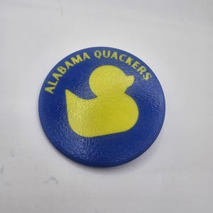 Puede incluir: Un objeto redondo azul y amarillo con las palabras "ALABAMA QUACKERS" en texto amarillo alrededor del borde. El centro presenta la forma de un pato amarillo. El objeto es probablemente un botón o similar.