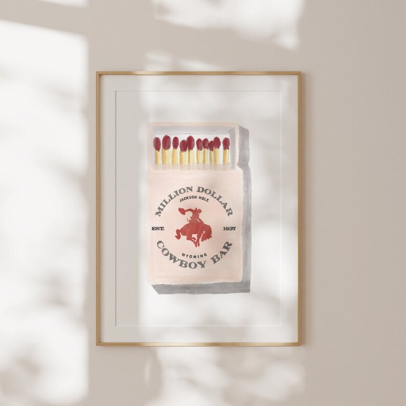 May include: A vintage-style illustration of a matchbox with a red and white design. The matchbox is labeled "Million Dollar Cowboy Bar" with the text "Jackson Hole, Wyoming, Est. 1937" printed on the front.