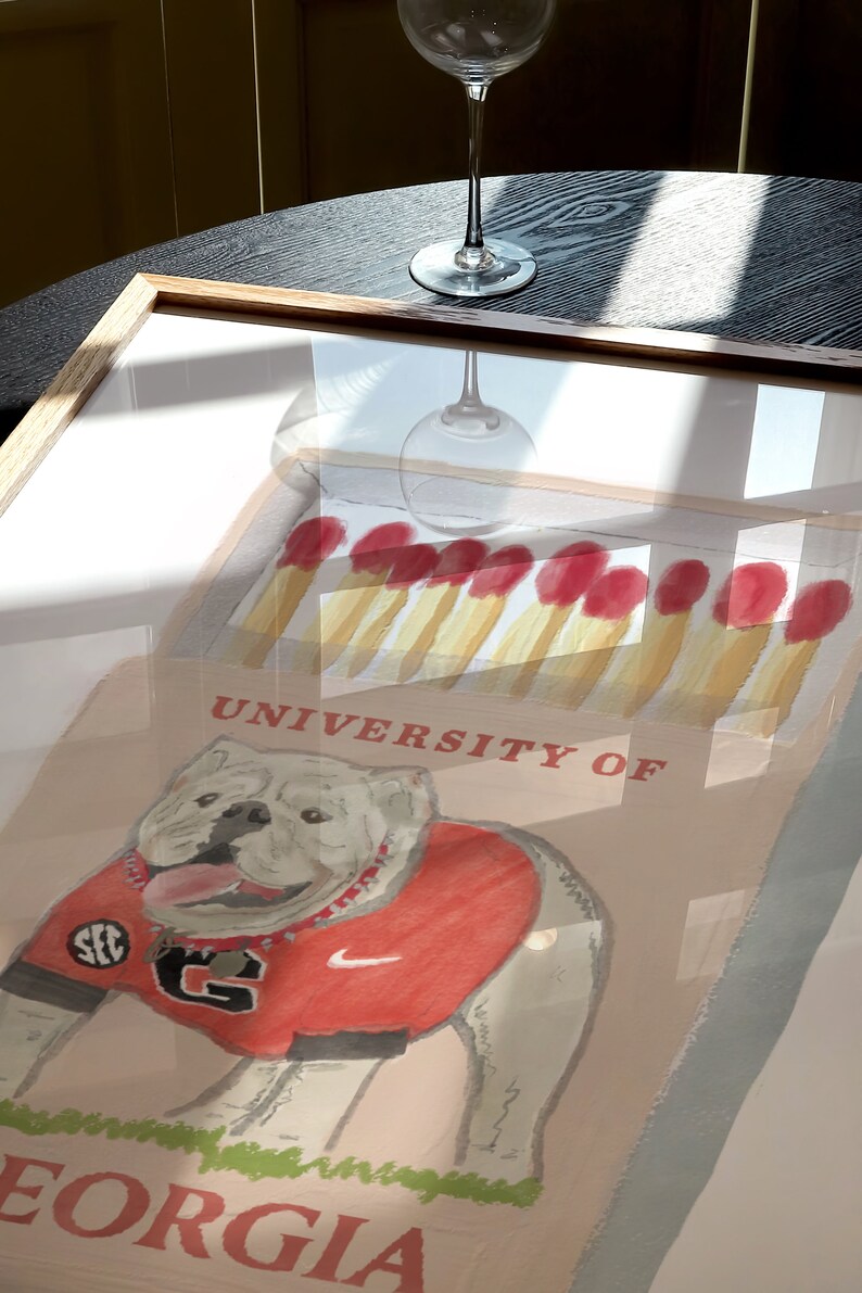 May include: A framed illustration of a white bulldog wearing a red jersey with the text "SEC" and "G" on it. The bulldog is standing on a box with the text "University of Georgia" on it. The box is filled with red-tipped matches.