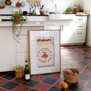 May include: A framed print of a vintage matchbox with the text "Million Dollar Cowboy Bar" and a red illustration of a cowboy riding a bucking horse. The print is hanging on a white wall in a kitchen.