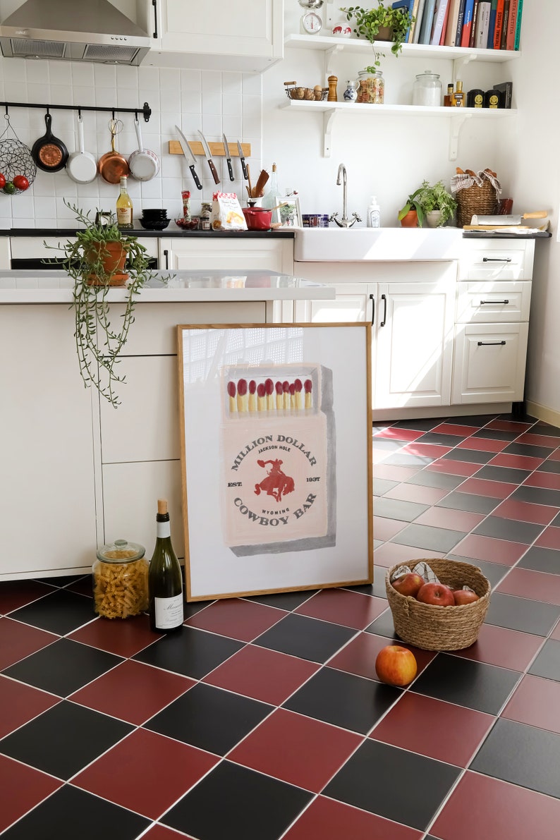 May include: A framed print of a vintage matchbox with the text "Million Dollar Cowboy Bar" and a red illustration of a cowboy riding a bucking horse. The print is hanging on a white wall in a kitchen.