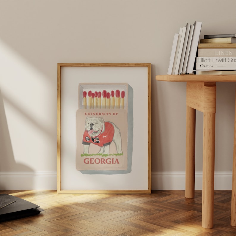 May include: A vintage-style illustration of a matchbox featuring a white bulldog wearing a red and black jersey with the text "UNIVERSITY OF GEORGIA". The bulldog is standing on a green patch of grass. The matchbox is filled with red-tipped matches.