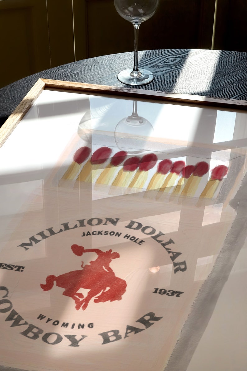 May include: A framed print of a vintage poster for the Million Dollar Cowboy Bar in Jackson Hole, Wyoming. The poster features a red cowboy riding a bucking horse with the text "Million Dollar Cowboy Bar" and "Jackson Hole, Wyoming, Est. 1937".
