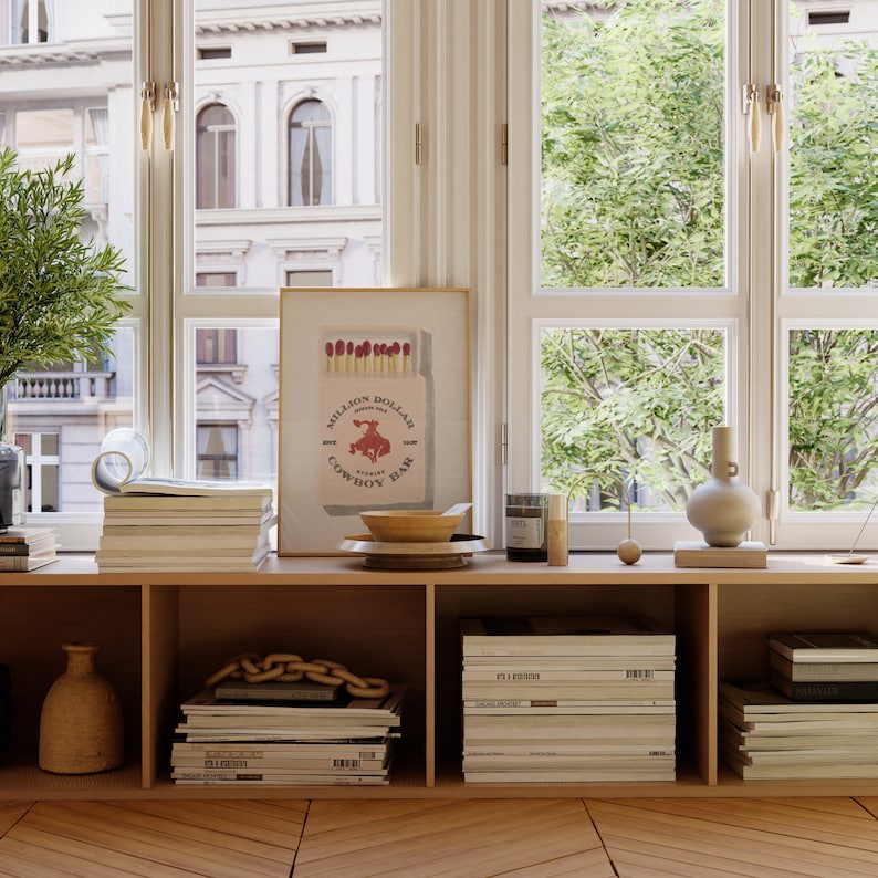 May include: A wooden shelf unit with a framed print of a red bull riding a cowboy on a box of matches. The print reads "Million Dollar Cowboy Bar". The shelf is decorated with a bowl, a vase, a candle, and a stack of magazines.