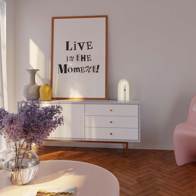 May include: A white dresser with four drawers and gold knobs. A framed print with the text "Live in the Moment!" hangs above the dresser. A white arch-shaped lamp with a gold accent sits on the dresser. A vase with purple flowers sits on a pink table in front of the dresser.