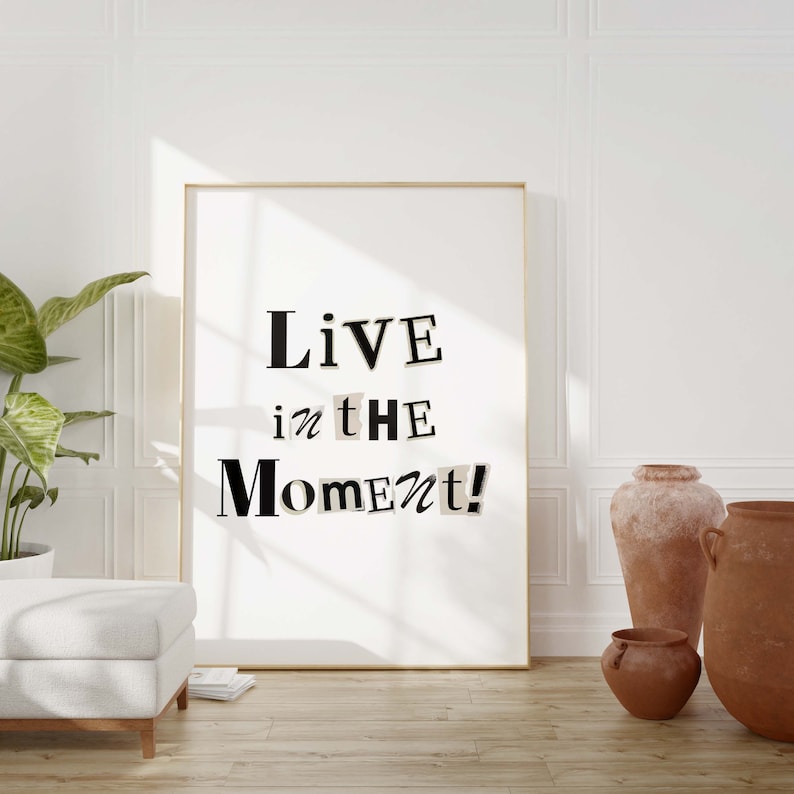 May include: A framed print with a white background and black text that reads "Live in the Moment!" The print is hanging on a white wall in a room with a potted plant and a wooden floor.