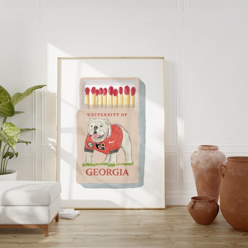May include: A vintage-style matchbox illustration featuring a white bulldog wearing a red and black University of Georgia jersey. The matchbox reads "University of Georgia" and has a red and white striped matchstick design.