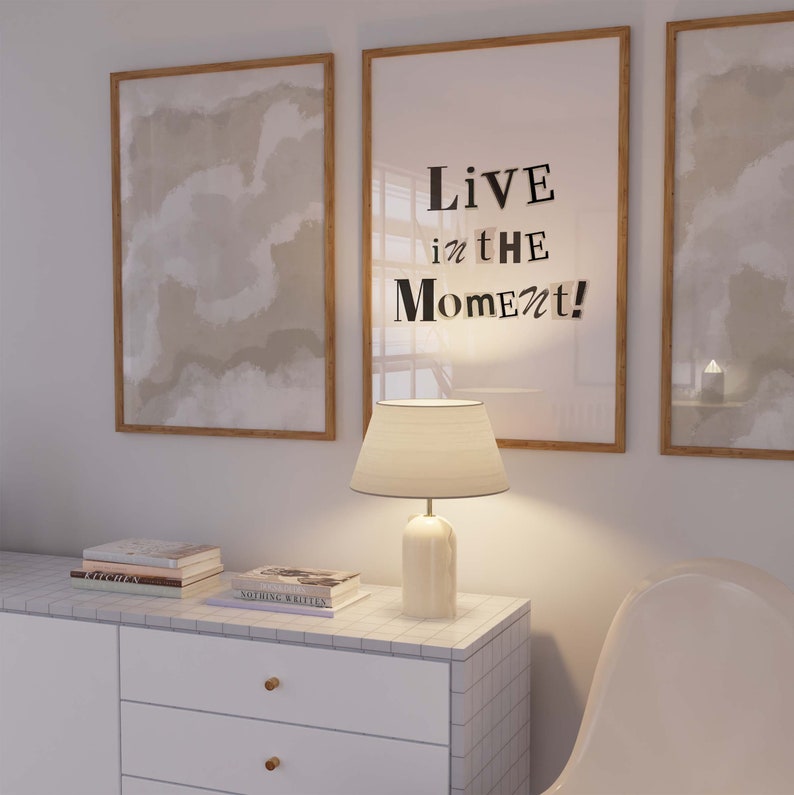 May include: A white lamp with a cream-colored shade sits on a white dresser with a tiled top. The dresser has two drawers. Two framed pictures hang on the wall behind the lamp. The picture on the right has the text "Live in the Moment!" printed on it.