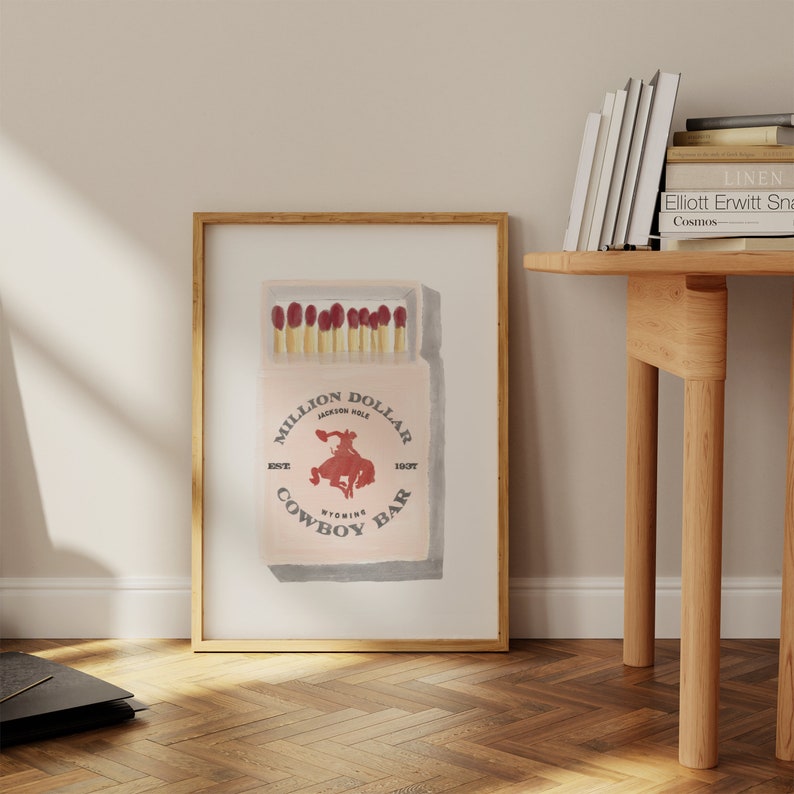 May include: A vintage-style illustration of a matchbox with the text "Million Dollar Cowboy Bar" and the location "Jackson Hole, Wyoming". The matchbox is illustrated in a watercolor style with red and white accents.